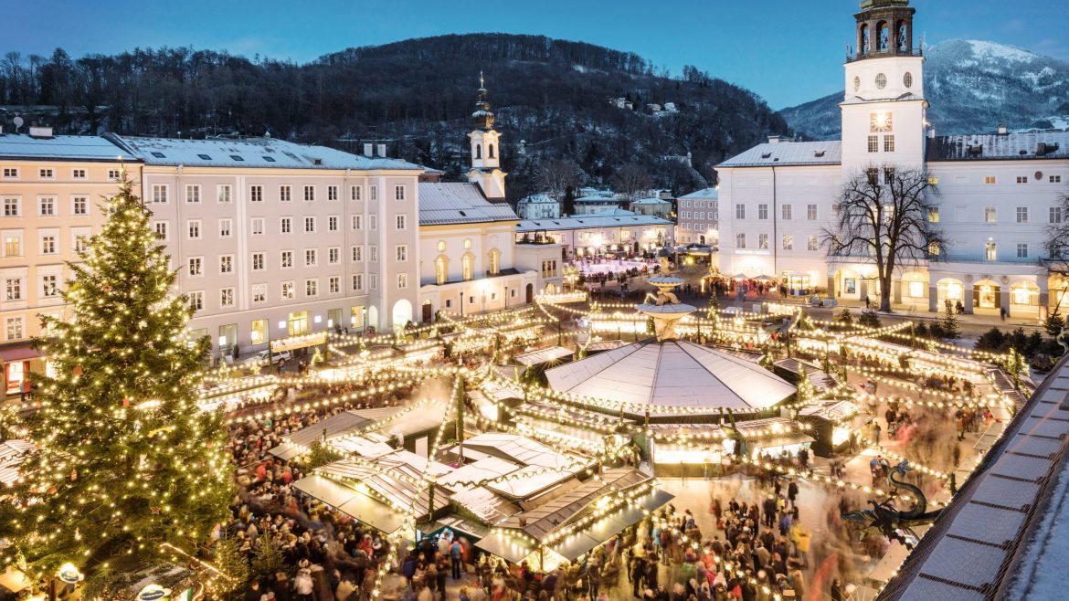 Ein festlicher Weihnachtsmarkt in der Abenddämmerung mit Lichtern, einem großen Baum und vielen Menschen auf einem verschneiten Stadtplatz.