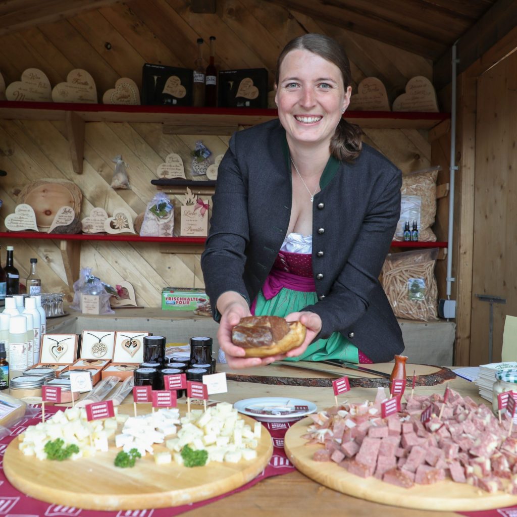 Lächelnde Frau an einem Marktstand, die Lebensmittel anbietet, mit Käse, Fleisch und Gläsern vor sich.