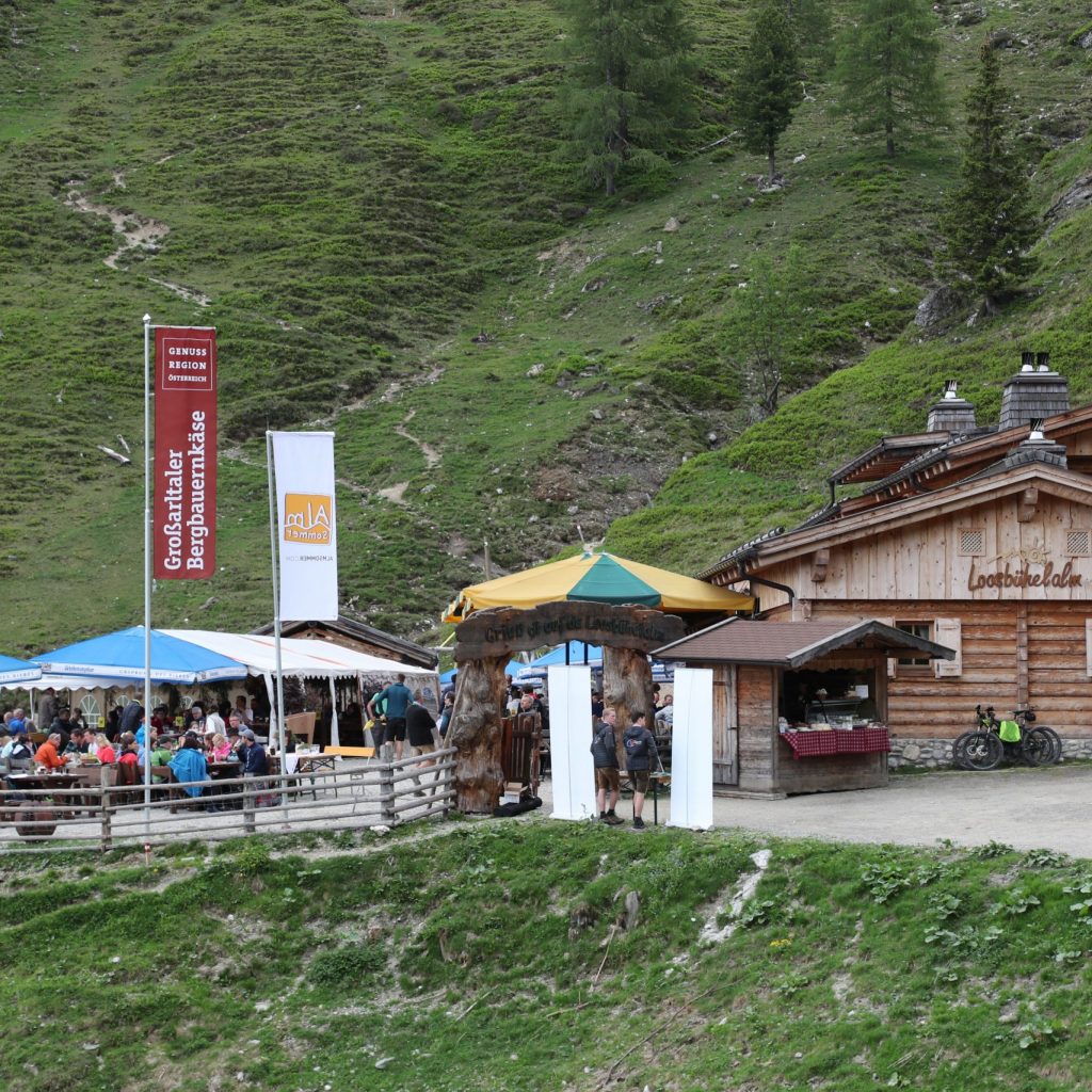 Menschen, die unter freiem Himmel in einem Berghüttencafé mit Sonnenschirmen speisen, umgeben von grasbewachsenen Hügeln und Bäumen.