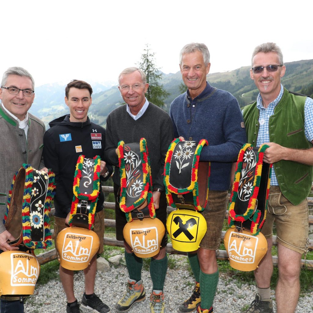 Fünf Männer in alpiner Kleidung halten geschmückte Kuhglocken und gelbe Helme in der Hand und lächeln in die Kamera.