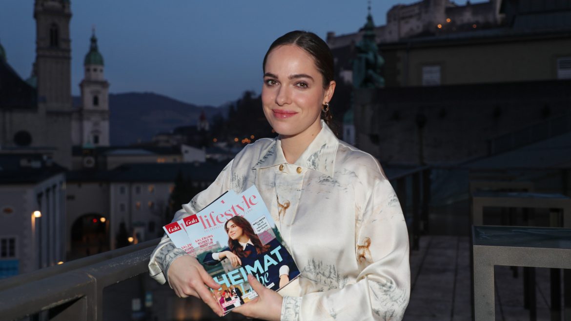 Frau, die in der Abenddämmerung auf einem Balkon steht und zwei Lifestyle-Magazine in der Hand hält, mit einer Stadtlandschaft im Hintergrund.