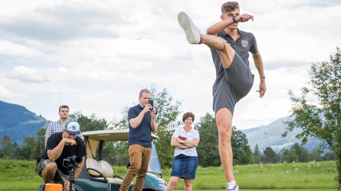 Ein Mann kickt hoch auf einem Golfplatz, während andere zusehen und filmen, mit einem Golfwagen und Bäumen im Hintergrund.