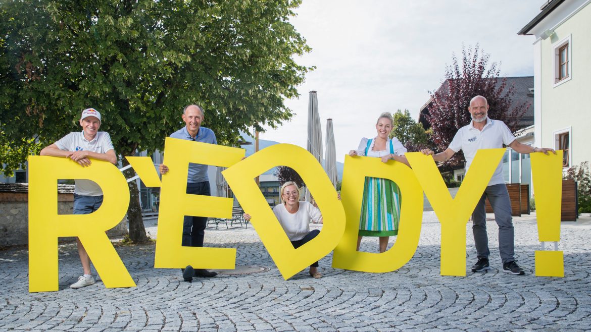 Fünf Personen stehen im Freien und halten große gelbe Buchstaben mit der Aufschrift REDDY! auf einer Kopfsteinpflasterstraße.