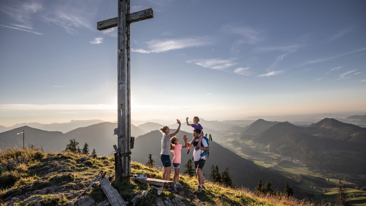 Eine Familie feiert bei einem großen Holzkreuz auf einem Berggipfel bei Sonnenuntergang mit Blick auf ein malerisches Tal.