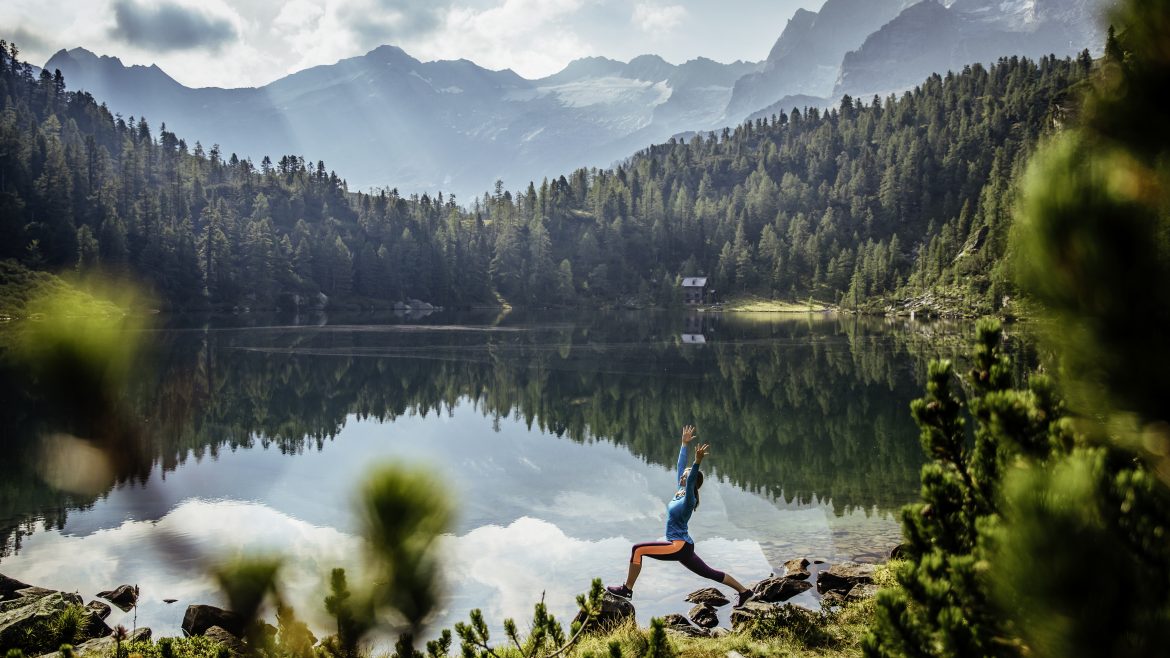 Person beim Yoga an einem ruhigen Bergsee, umgeben von Bäumen und fernen Gipfeln unter einem bewölkten Himmel.