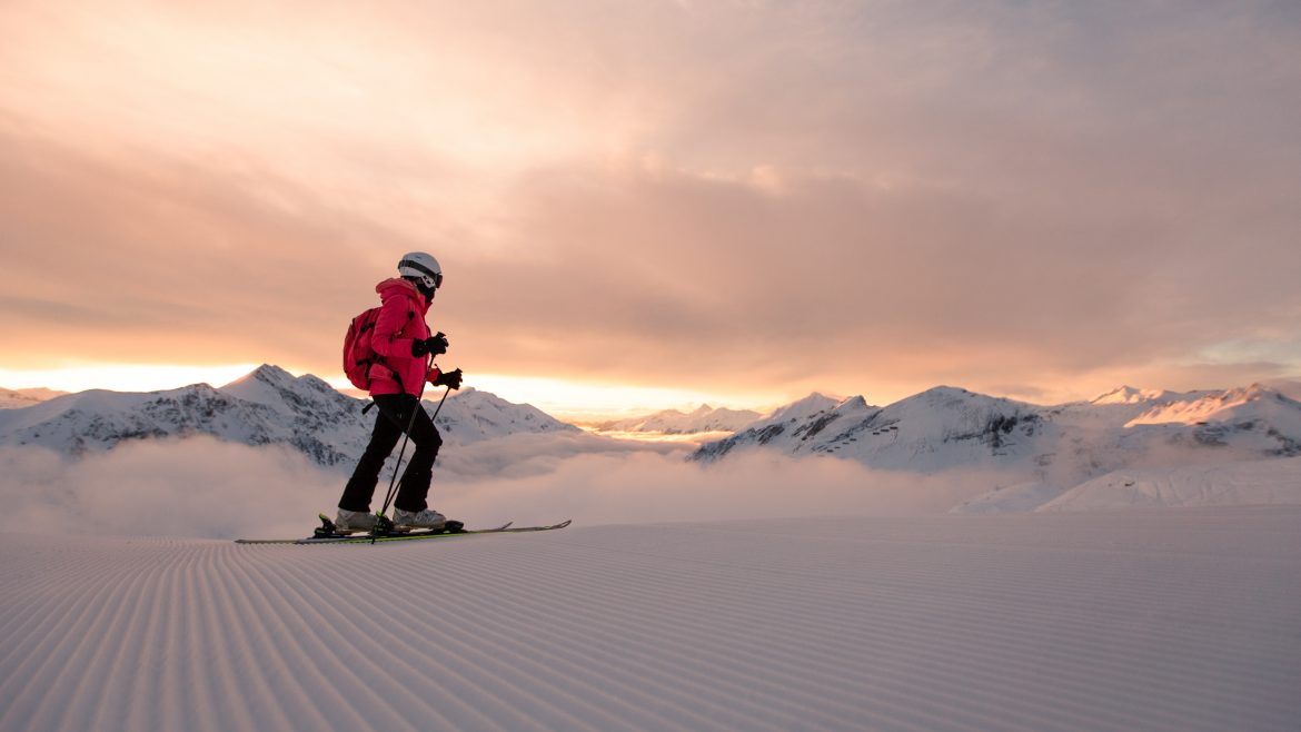 Person beim Skifahren bei Sonnenaufgang auf einer präparierten Schneepiste, mit Bergen und Wolken im Hintergrund.