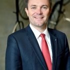 Ein Mann in einem dunklen Anzug mit roter Krawatte steht lächelnd in einem Haus mit alten Fahrrädern im Hintergrund.
