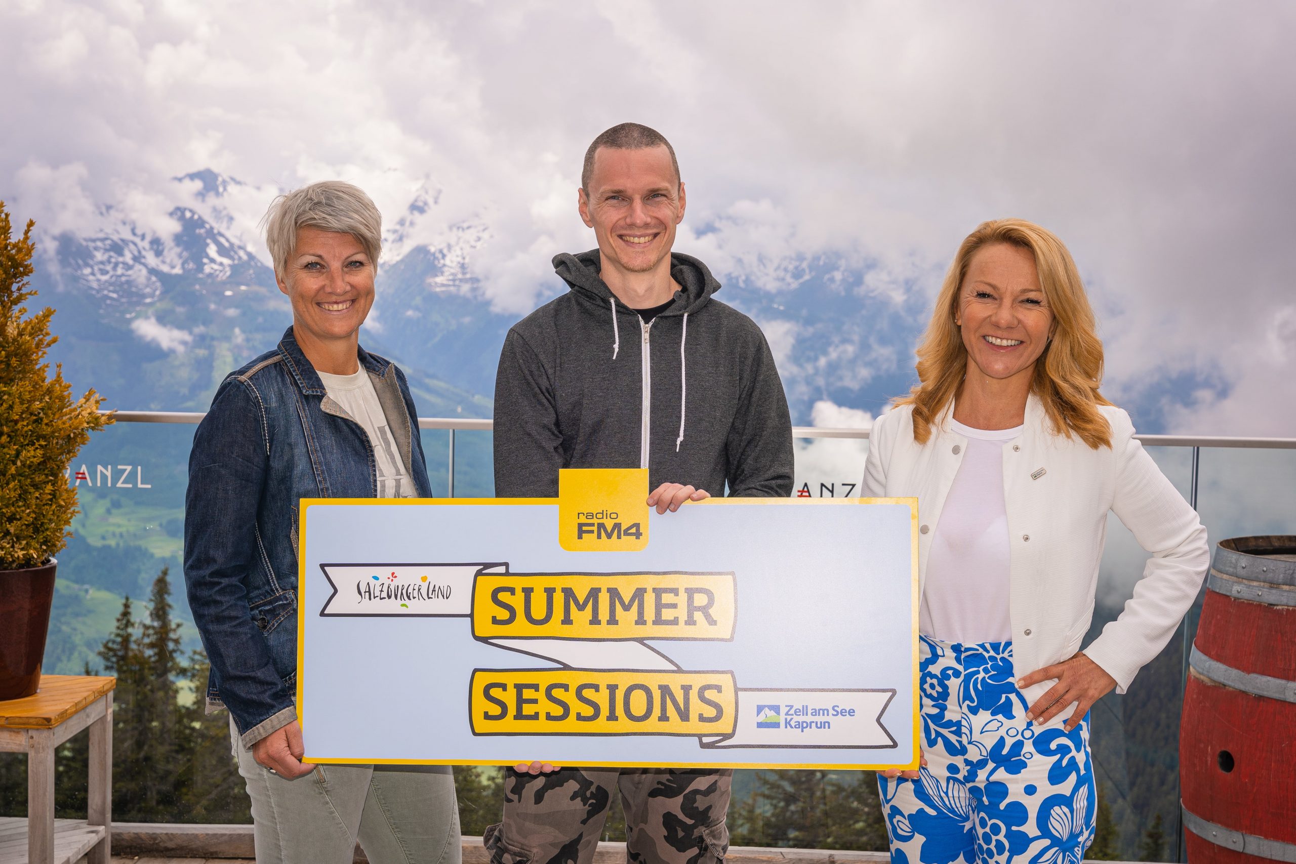 Drei Personen lächeln und halten ein Summer Sessions Schild mit Bergen und bewölktem Himmel im Hintergrund.