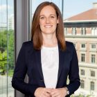 Eine Frau mit glattem braunem Haar in einem marineblauen Blazer lächelt, während sie an einem Fenster steht, aus dem die Gebäude der Stadt zu sehen sind.