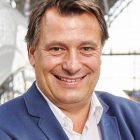 Lächelnder Mann in blauer Anzugsjacke und weißem Hemd steht in einem Haus mit unscharfem Hintergrund.