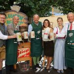 Sechs Personen in traditioneller Kleidung lächeln und halten Bierkrüge bei einem Bierfest im Freien.