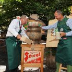 Zwei Männer in Schürzen zapfen im Freien ein hölzernes Bierfass an, während ein weiterer Mann lächelnd und mit Bierkrügen in der Hand zusieht.