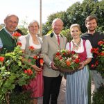 Fünf Personen in traditioneller österreichischer Kleidung stehen im Freien, lächeln und halten Blumen und einen Korb mit Gemüse.
