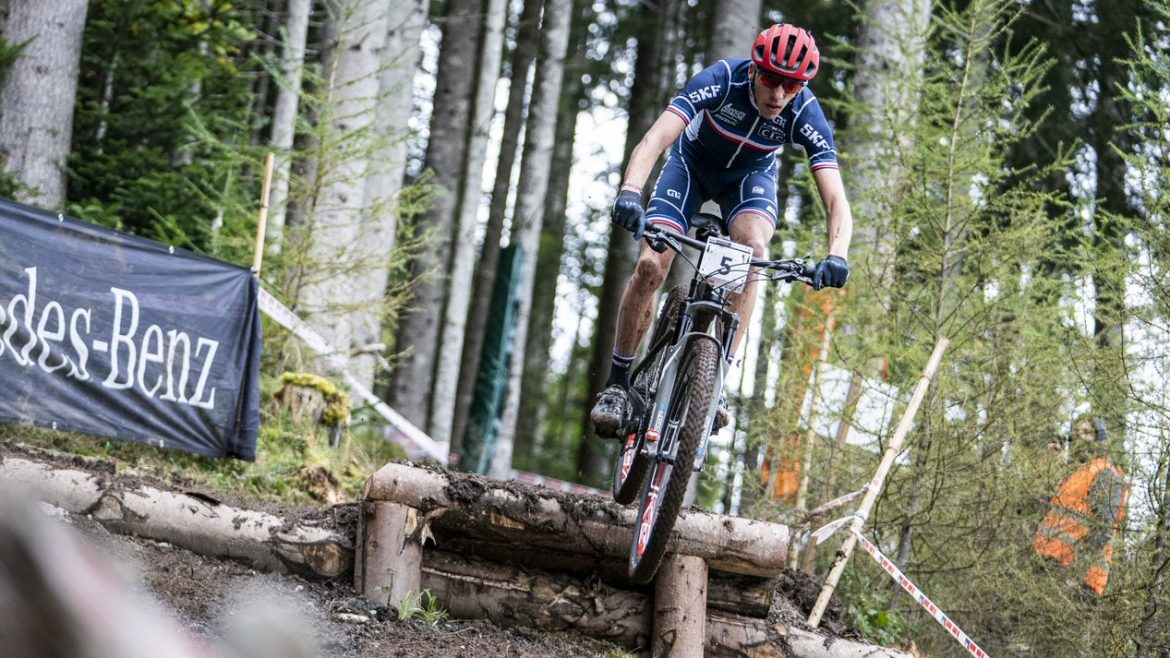 Ein Radfahrer in blauer Montur fährt mit seinem Mountainbike über ein Hindernis aus Baumstämmen bei einem Rennen auf einem Waldweg.