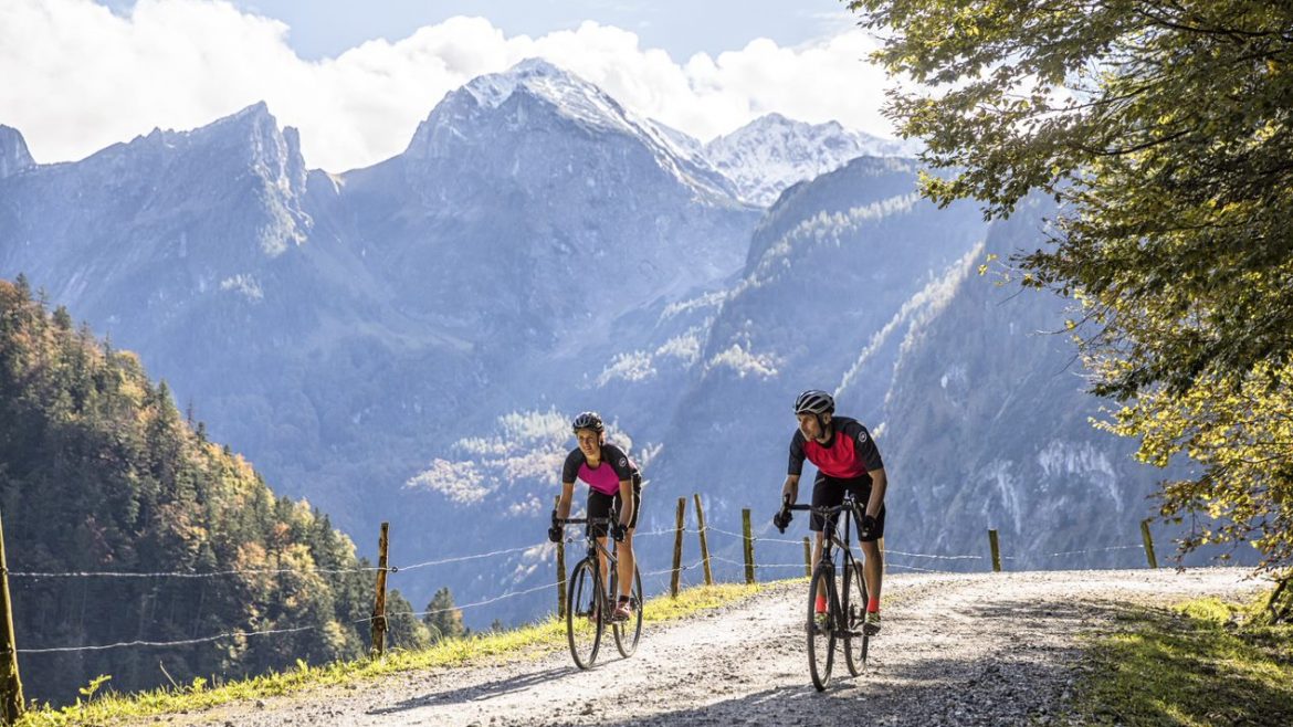 Zwei Radfahrer fahren an einem sonnigen Tag auf einem Schotterweg mit Bergen und Bäumen im Hintergrund.