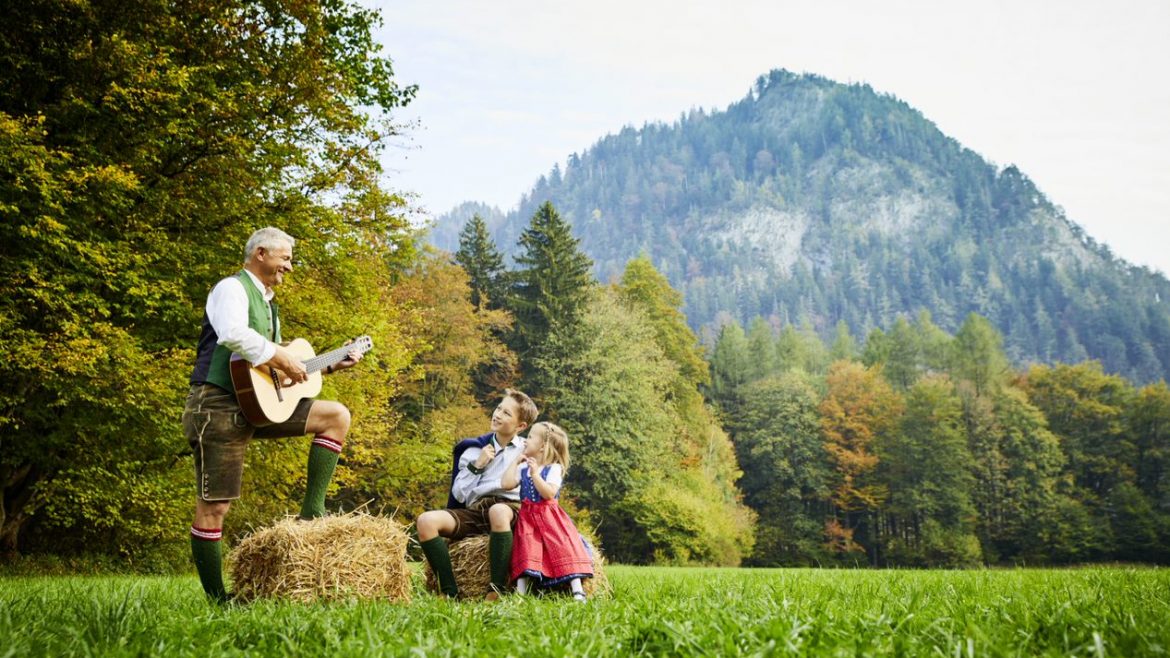 Ein älterer Mann spielt Gitarre für zwei Kinder auf einer Wiese mit Bergen und Bäumen im Hintergrund.