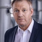 Ein Mann in einem marineblauen Anzug und weißem Hemd steht in einem Haus und schaut direkt in die Kamera.
