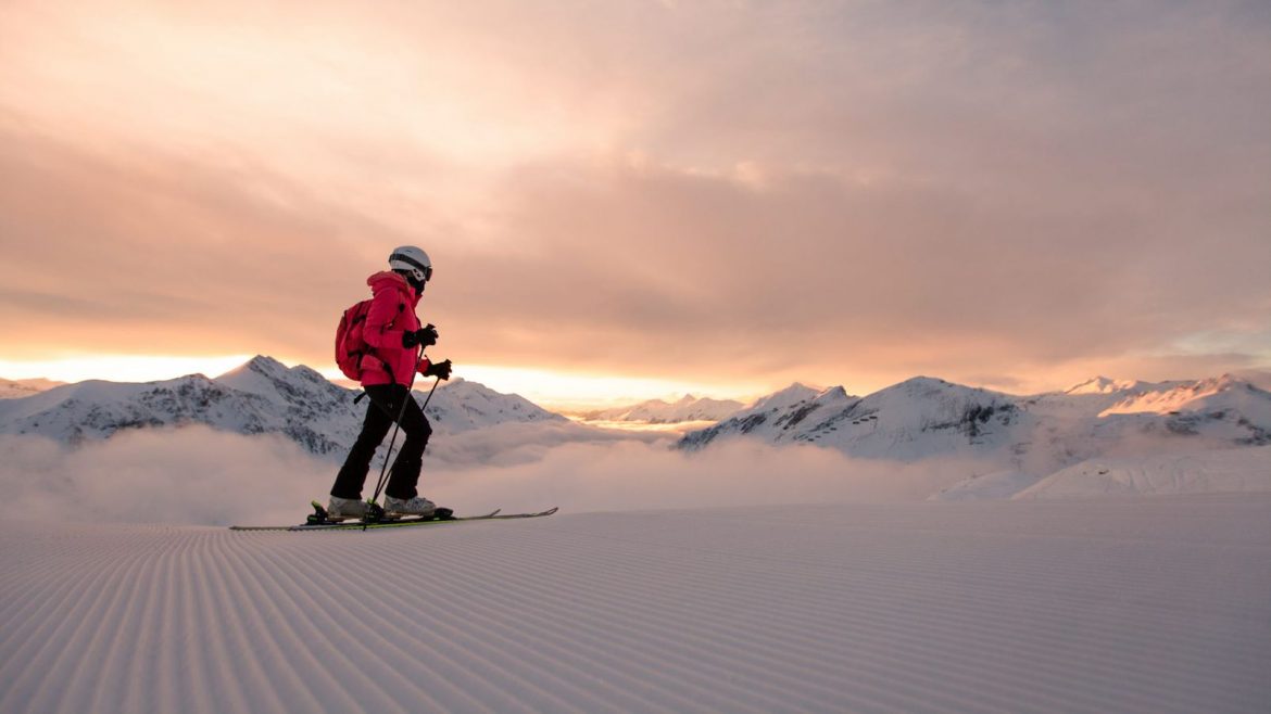 Ein Skifahrer in rosafarbener Kleidung gleitet bei Sonnenaufgang über eine verschneite Piste, mit Bergen und Wolken im Hintergrund.