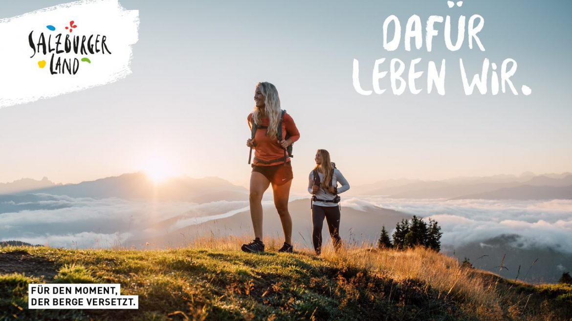 Zwei Frauen wandern bei Sonnenaufgang auf einem grasbewachsenen Berg mit Wolken und Gipfeln im Hintergrund.