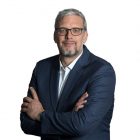 Mann mit grauen Haaren und Brille in einem dunklen Anzug, stehend mit verschränkten Armen vor einem weißen Hintergrund.
