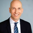 Glatzköpfiger Mann in Anzug und gestreifter Krawatte, der vor einem schlichten hellblauen Hintergrund lächelt.