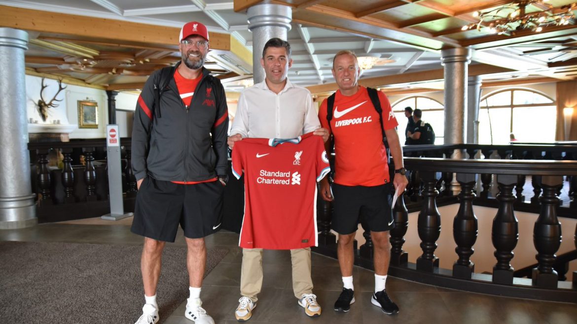 Drei Männer posieren in einem Innenraum; einer hält ein Trikot des FC Liverpool. Zwei tragen Liverpooler Sportkleidung und stehen auf beiden Seiten.