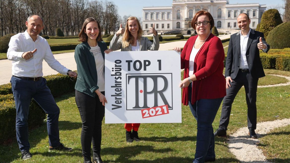 Fünf lächelnde Menschen posieren im Freien und halten ein Schild mit der Aufschrift Verkehrsbüro TOP 1 TPR 2021, dahinter ein großes Gebäude.