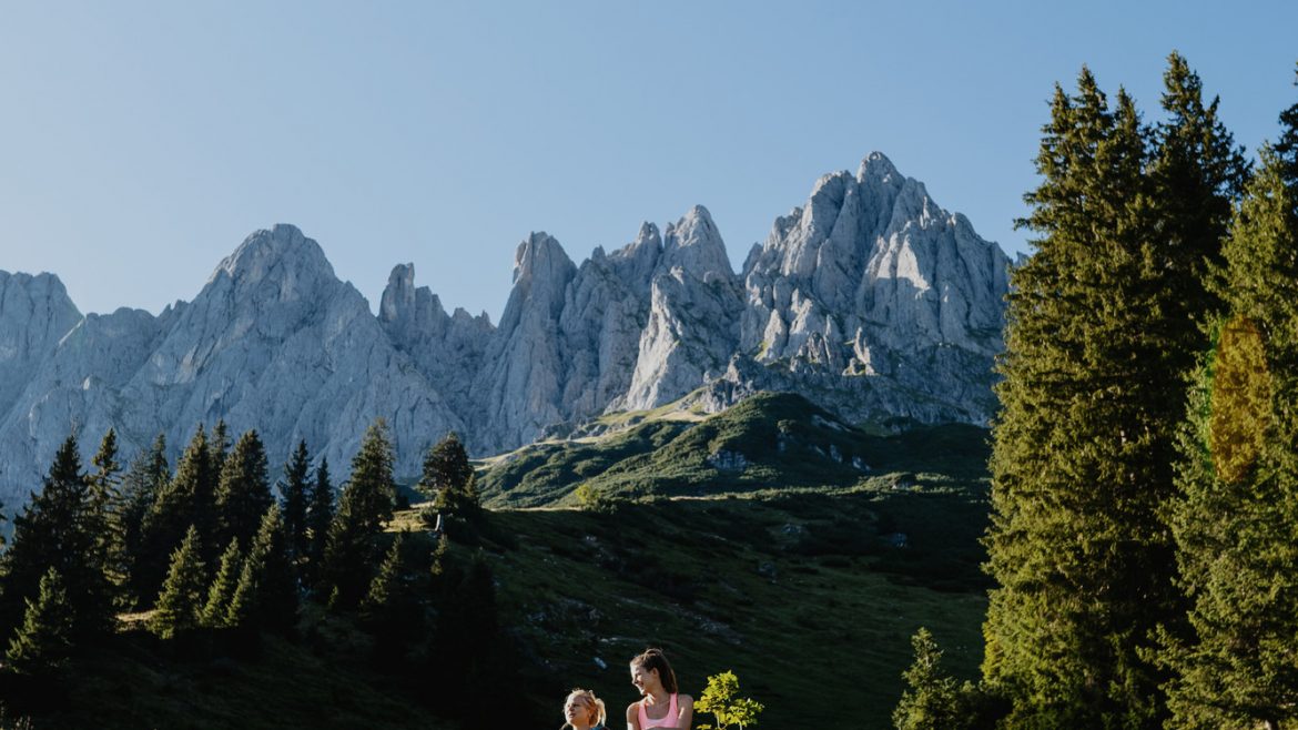 Zwei Personen sitzen auf Felsen in einer grasbewachsenen Berglandschaft mit hohen Kiefern und felsigen Gipfeln im Hintergrund.