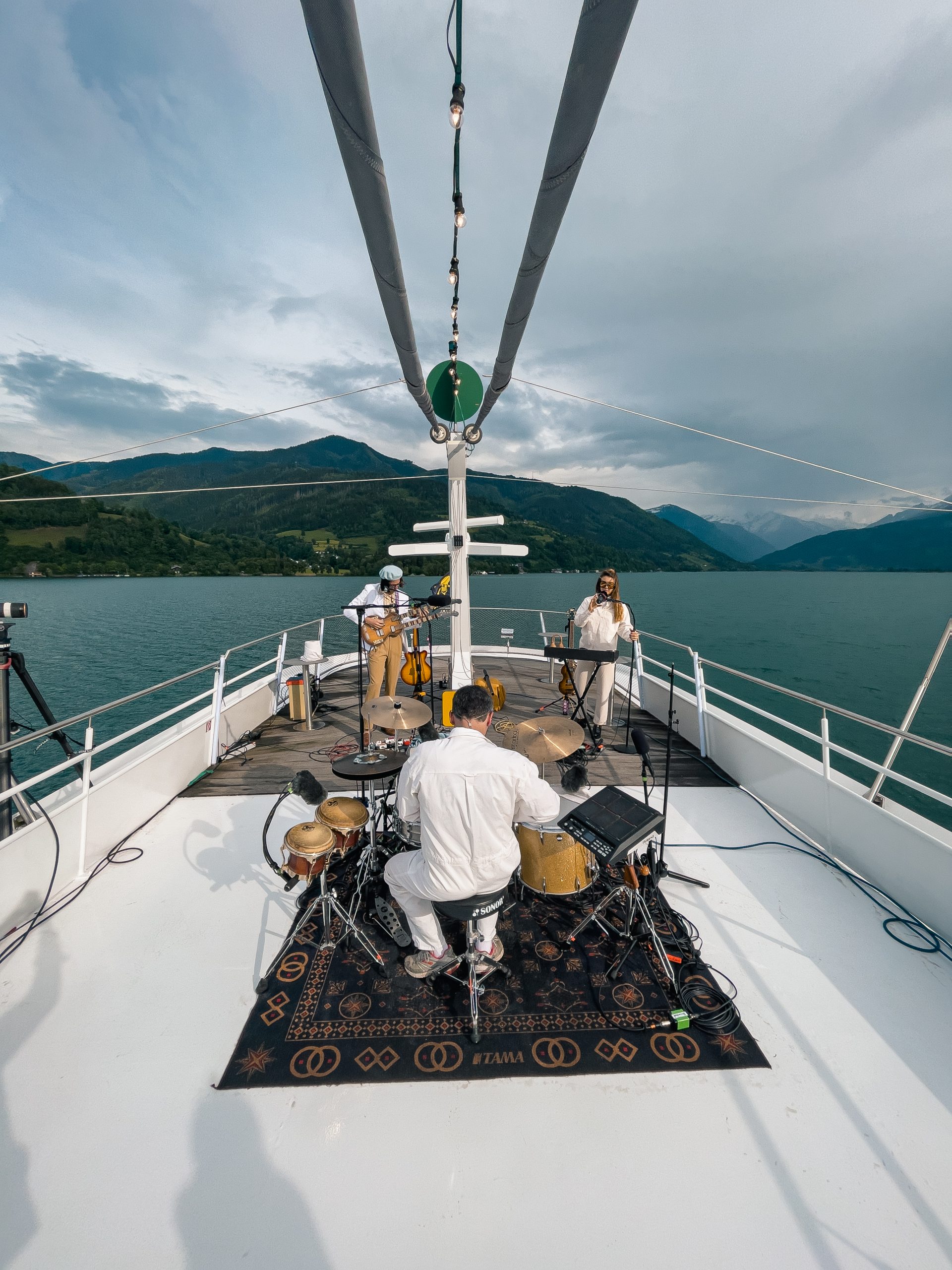 Eine Band tritt auf einem Bootsdeck auf, umgeben von einem See und Bergen unter einem bewölkten Himmel.