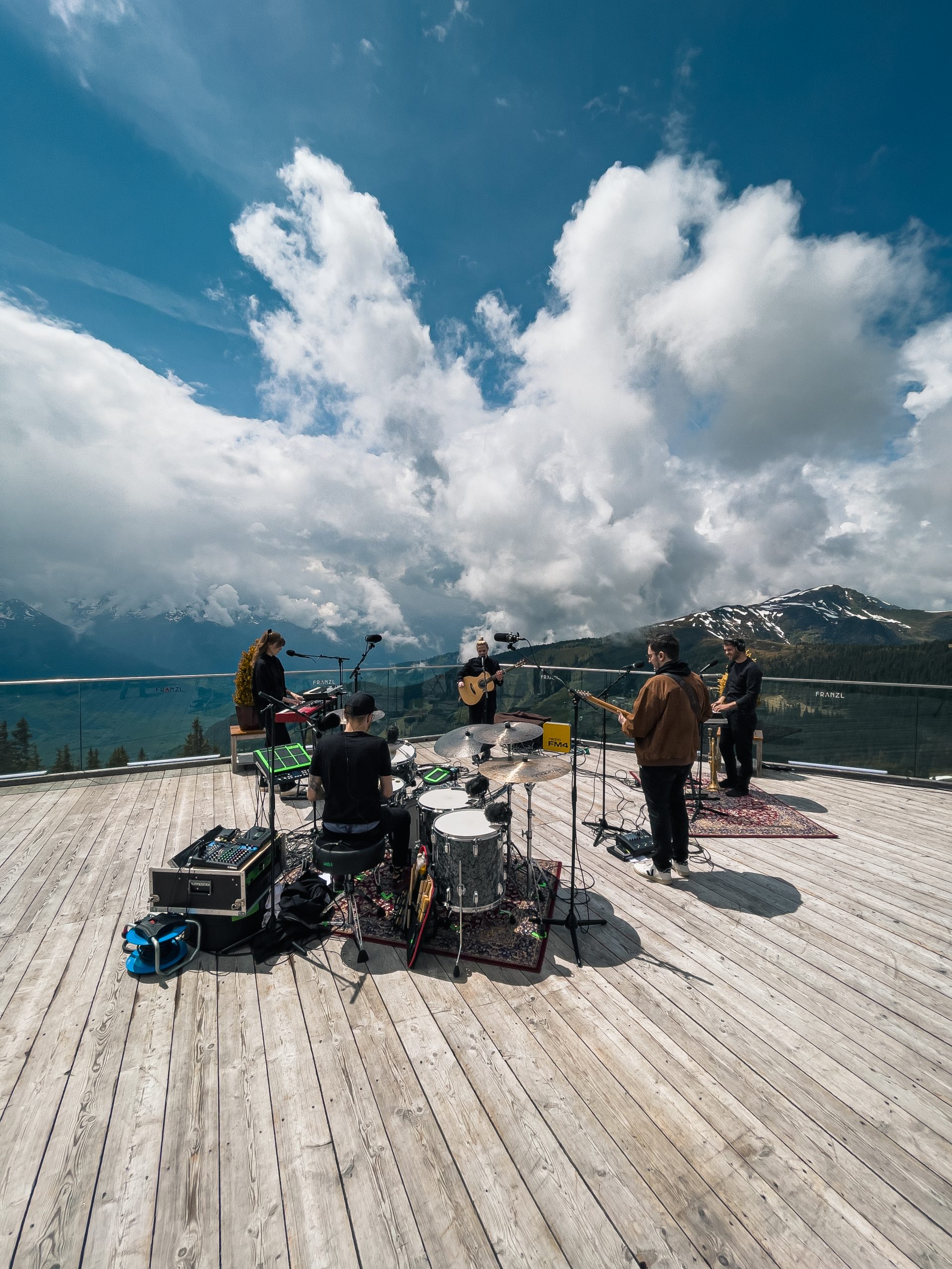 Eine Band spielt auf einer Holzterrasse im Freien mit Bergen und Wolken im Hintergrund.
