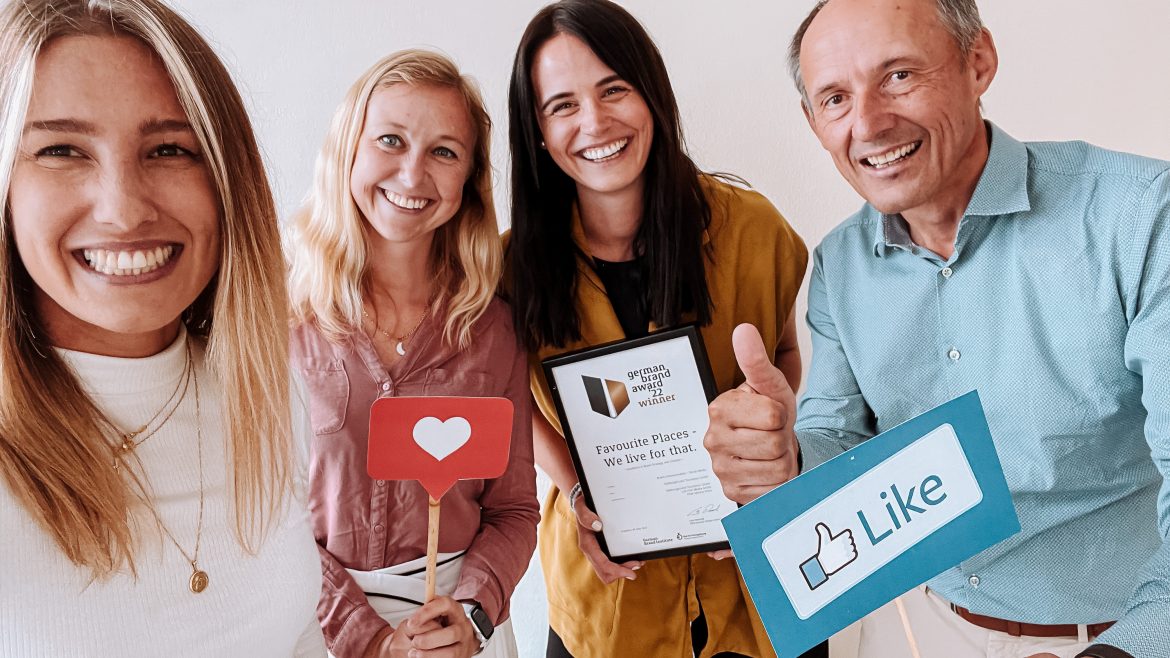 Vier lächelnde Menschen posieren mit einer Urkunde und Requisiten aus den sozialen Medien, darunter ein Herz und ein Like-Zeichen.