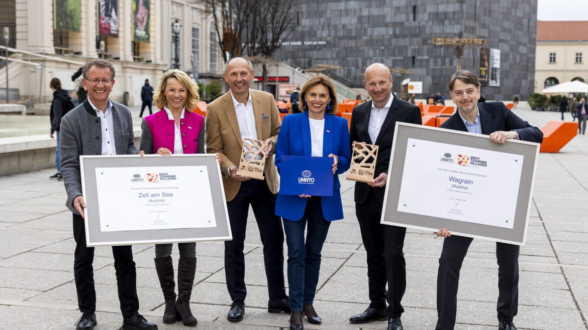 Sechs Personen stehen auf einem Stadtplatz mit Urkunden und Plaketten in der Hand und lächeln in die Kamera.