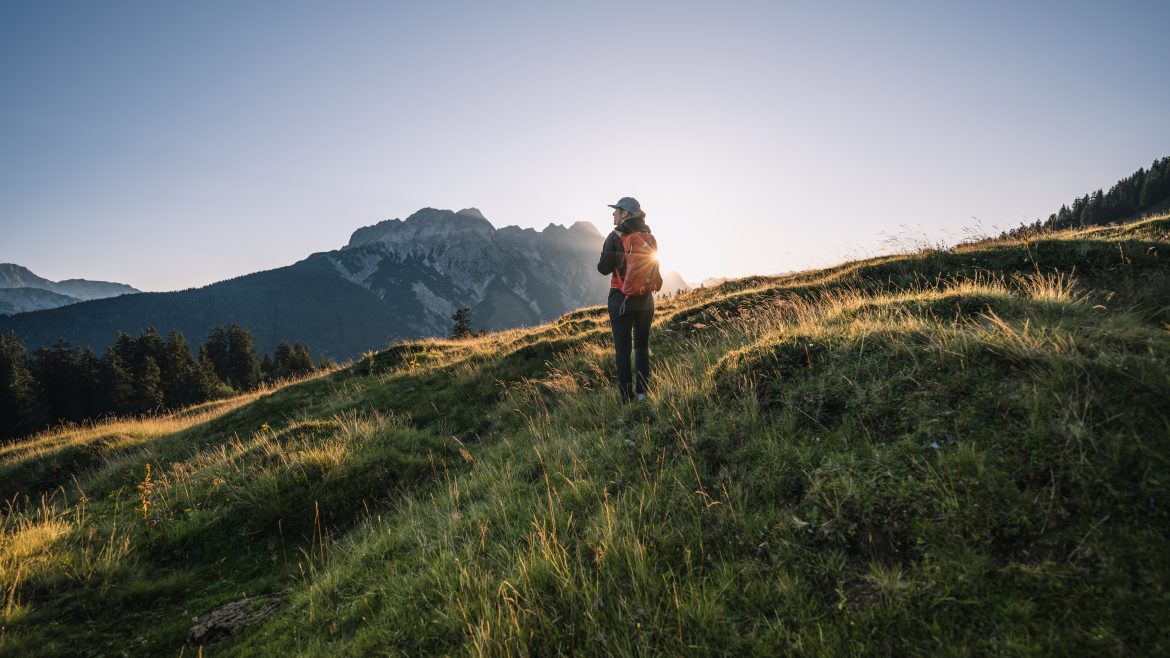 Person beim Wandern auf einem grasbewachsenen Hügel bei Sonnenaufgang mit Bergen und Bäumen im Hintergrund.
