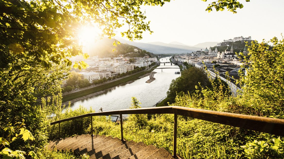 Steinstufen führen hinunter zu einem Fluss und einer Stadtlandschaft bei Sonnenuntergang, eingerahmt von grünen Blättern und fernen Hügeln.