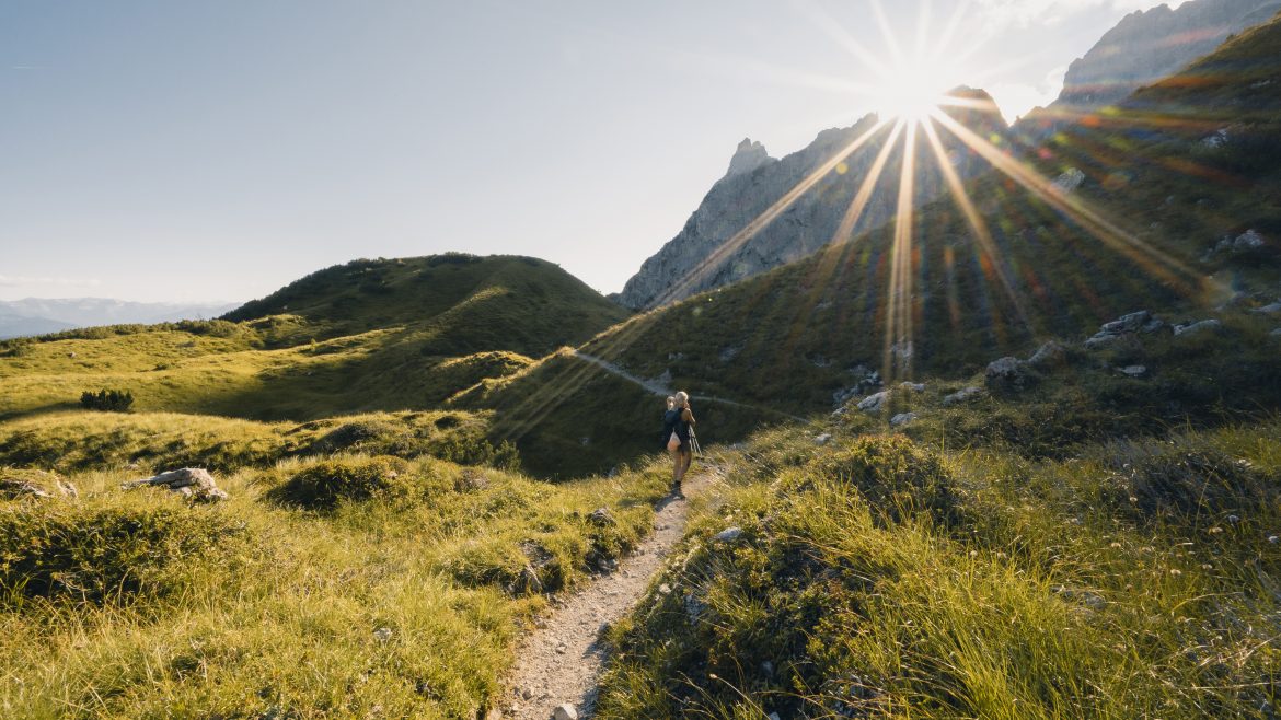 Eine Person wandert auf einem grasbewachsenen Bergpfad im hellen Sonnenlicht, wobei die Sonnenstrahlen über die entfernten Gipfel scheinen.