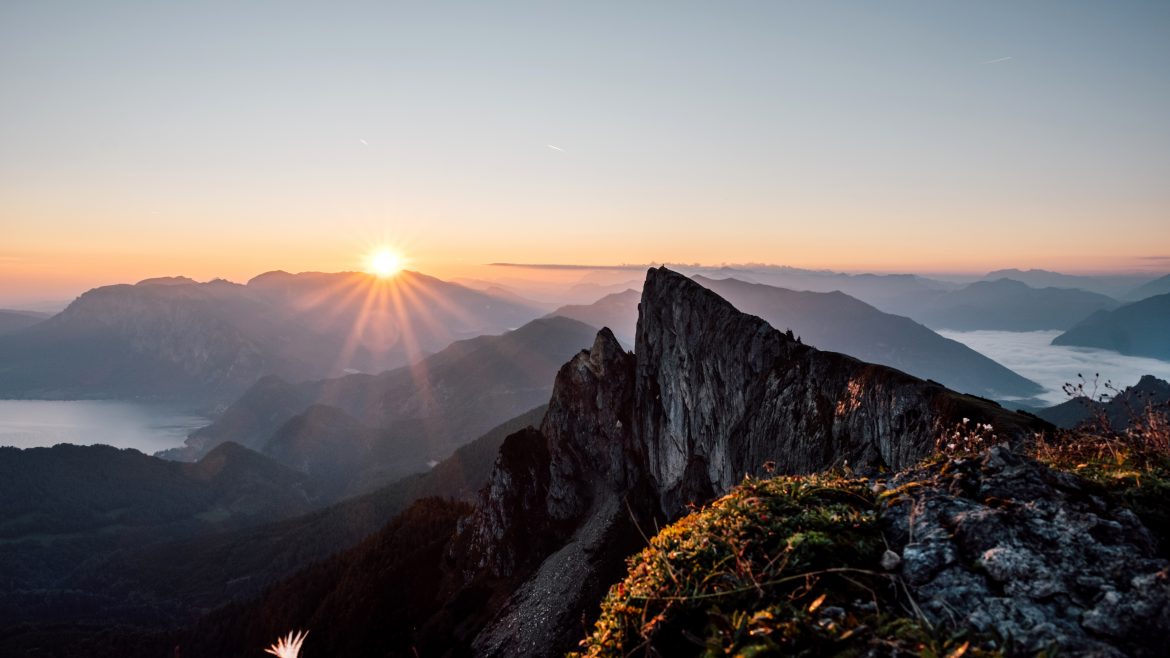 Sonnenaufgang über Berggipfeln mit felsigen Klippen und einem Tal darunter, getaucht in warmes Morgenlicht.