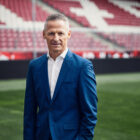 Ein Mann in einem blauen Anzug steht auf einem Fußballfeld mit leeren roten und weißen Stadionsitzen im Hintergrund.