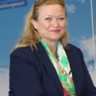 Eine Frau mit blondem Haar in einem marineblauen Blazer und einem bunten Schal, die vor einem blauen Hintergrund lächelt.