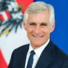 Lächelnder Mann im Anzug, im Hintergrund die Fahnen Österreichs und der Europäischen Union.