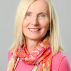 Lächelnde Frau mit blondem Haar, die ein rosa Oberteil und einen bunt gemusterten Schal trägt, vor einem hellgrauen Hintergrund.