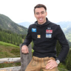 Ein Mann in Sportkleidung sitzt auf einem Holzzaun mit Bergen und grünen Hügeln im Hintergrund.