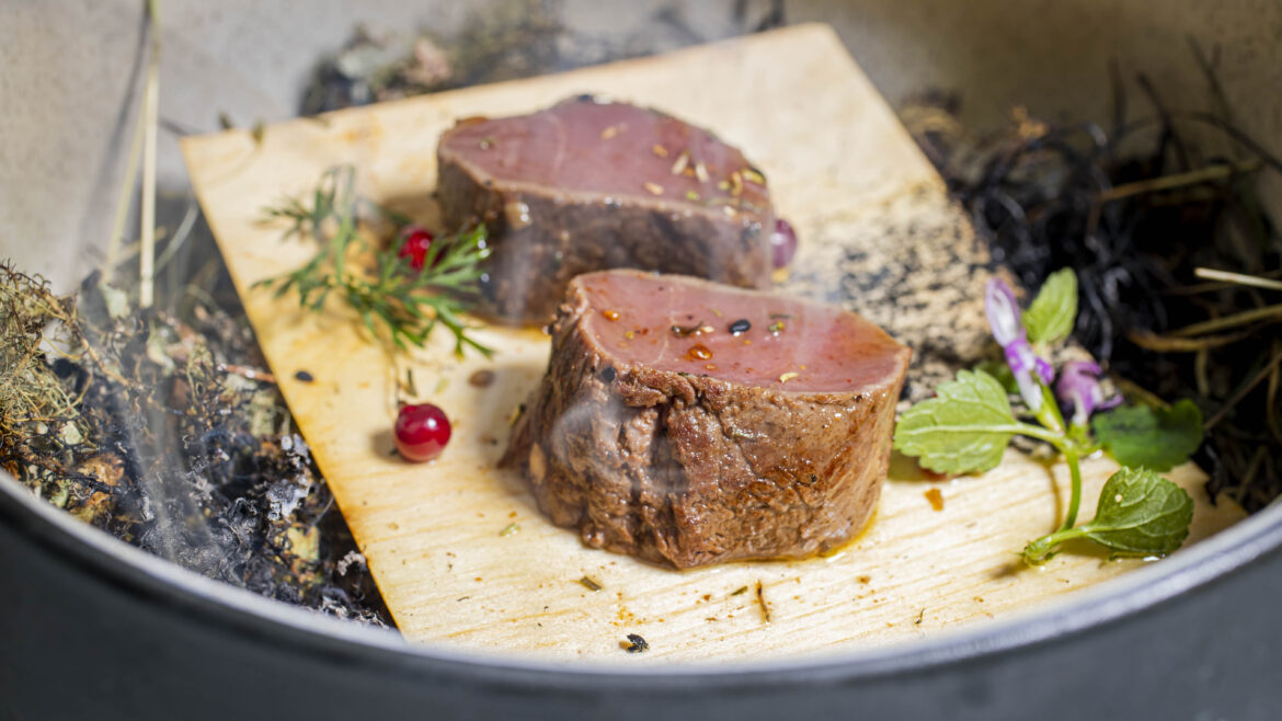 Zwei Scheiben gebratenes Steak auf einem Holzbrett, garniert mit Kräutern, Beeren und essbaren Blüten.