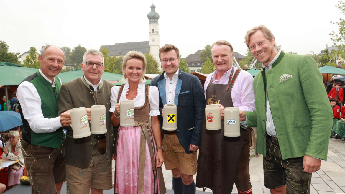 Sechs Personen in traditioneller österreichischer Kleidung halten Bierkrüge auf einem Fest im Freien mit einer Kirche im Hintergrund.