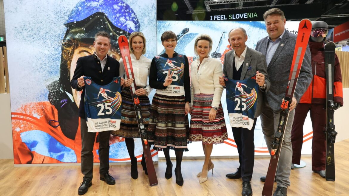 Sechs lächelnde Personen mit Skilätzchen und Skiern in Winterkleidung bei einer Indoor-Veranstaltung vor einer Skikulisse.