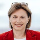 Lächelnde Frau mit kurzen braunen Haaren, Brille auf dem Kopf, in einem roten Blazer und weißem Oberteil, vor einem hellen Hintergrund.