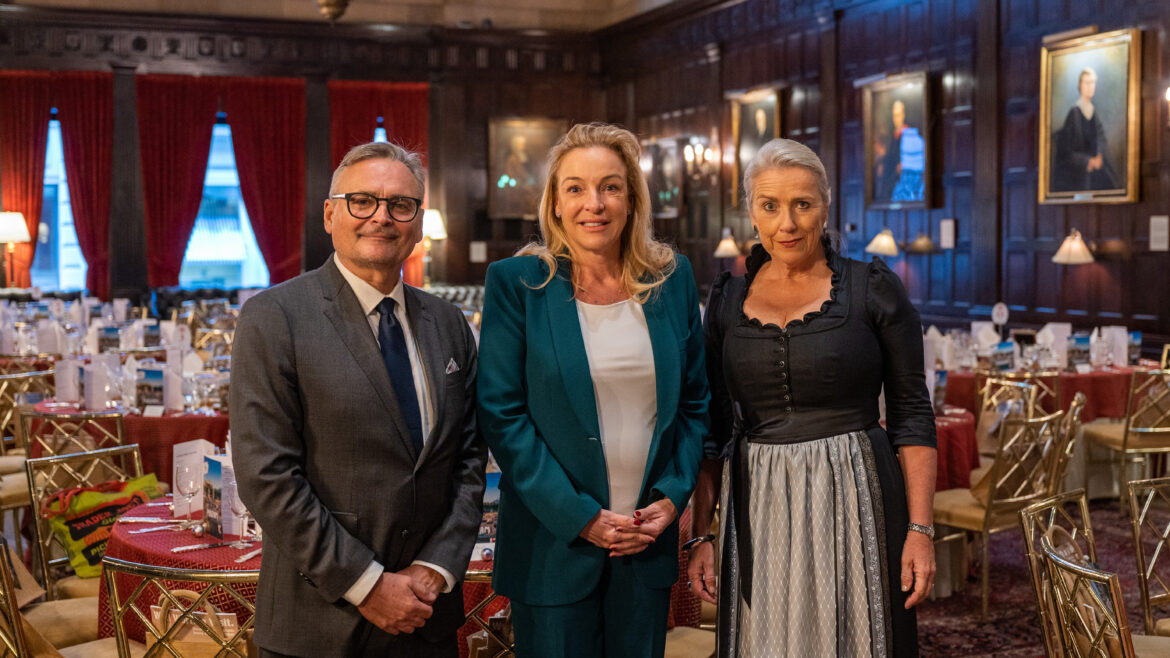 Drei formell gekleidete Personen stehen in einem eleganten, holzgetäfelten Speisesaal mit Tischen, die für eine Veranstaltung gedeckt sind.