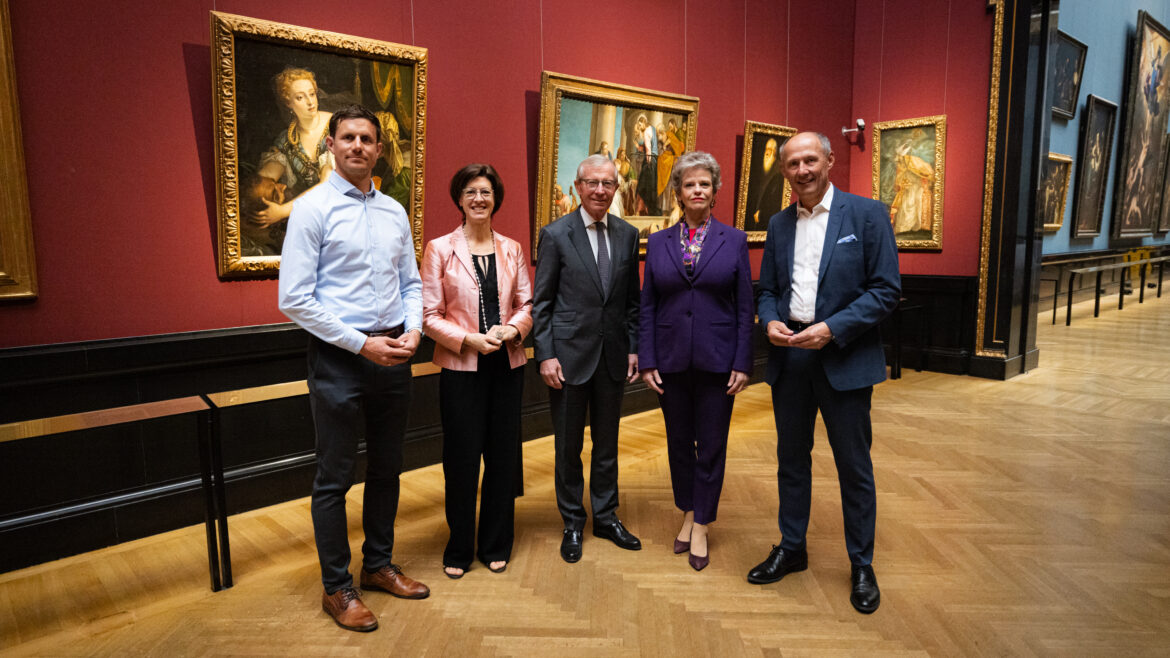 Fünf Personen stehen in einer Kunstgalerie mit gerahmten Gemälden an den roten Wänden hinter ihnen.