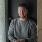 Ein Mann mit kurzen braunen Haaren und einem Bart lächelt, während er in Freizeitkleidung am Fenster sitzt.