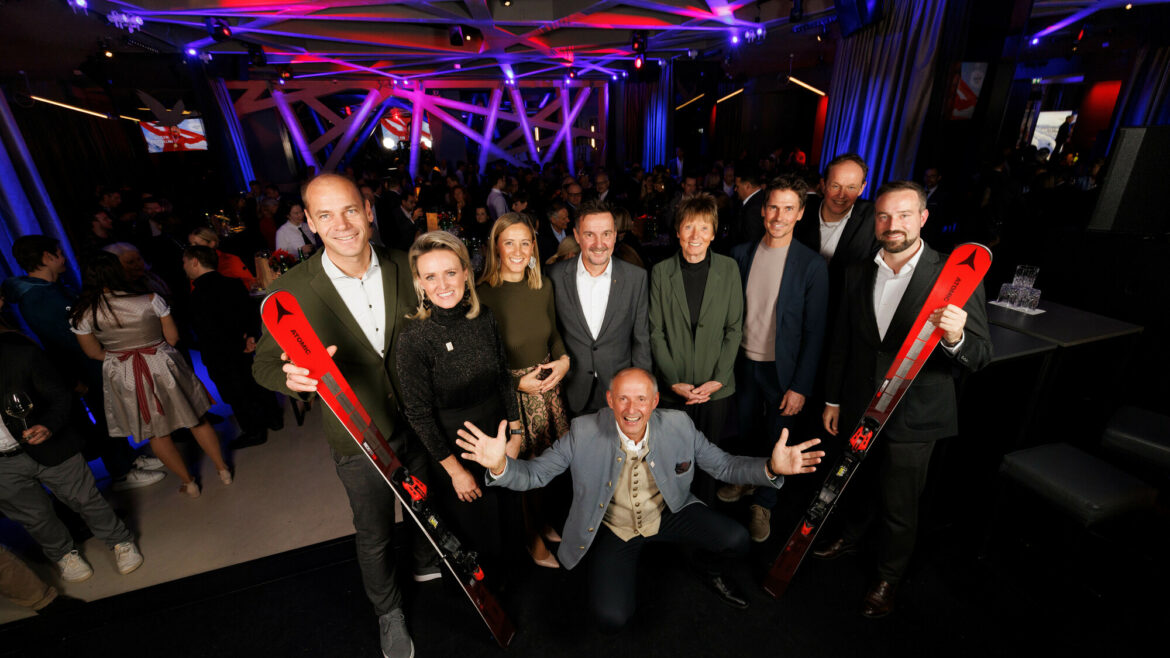 Eine Gruppe von Menschen posiert und lächelt bei einer Indoor-Veranstaltung, zwei halten rote Skier, im Hintergrund eine Menschenmenge.