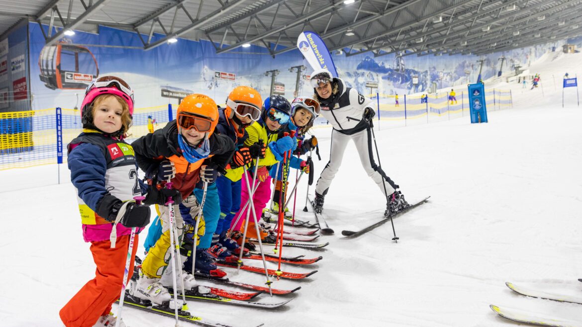 Ein Skilehrer posiert mit einer Gruppe von Kindern in farbenfroher Ausrüstung auf einer Indoor-Skipiste.